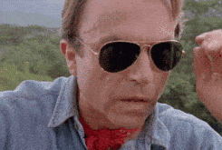 A man wearing sunglasses slowly removes them with a surprised or amazed expression on his face. He is dressed in a light blue denim shirt and a red bandana around his neck, with a green, scenic background behind him. - Choice Hacking Academy A man wearing sunglasses slowly removes them with a surprised or amazed expression on his face. He is dressed in a light blue denim shirt and a red bandana around his neck, with a green, scenic background behind him.