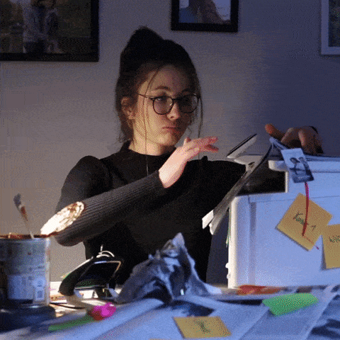 A woman with glasses and long hair in a bun is sitting at a cluttered desk, fanning herself with papers. The desk has various items, including sticky notes, a cup with pens, and crumpled paper. The room is dimly lit, and framed photos are visible on the wall behind her. - Choice Hacking Academy A woman with glasses and long hair in a bun is sitting at a cluttered desk, fanning herself with papers. The desk has various items, including sticky notes, a cup with pens, and crumpled paper. The room is dimly lit, and framed photos are visible on the wall behind her.