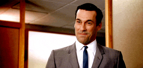 A man in a grey suit and blue tie is standing indoors with wood paneling and a glass window behind him. He has a slight smile on his face and raises his eyebrows in a suggestive or self-assured manner.