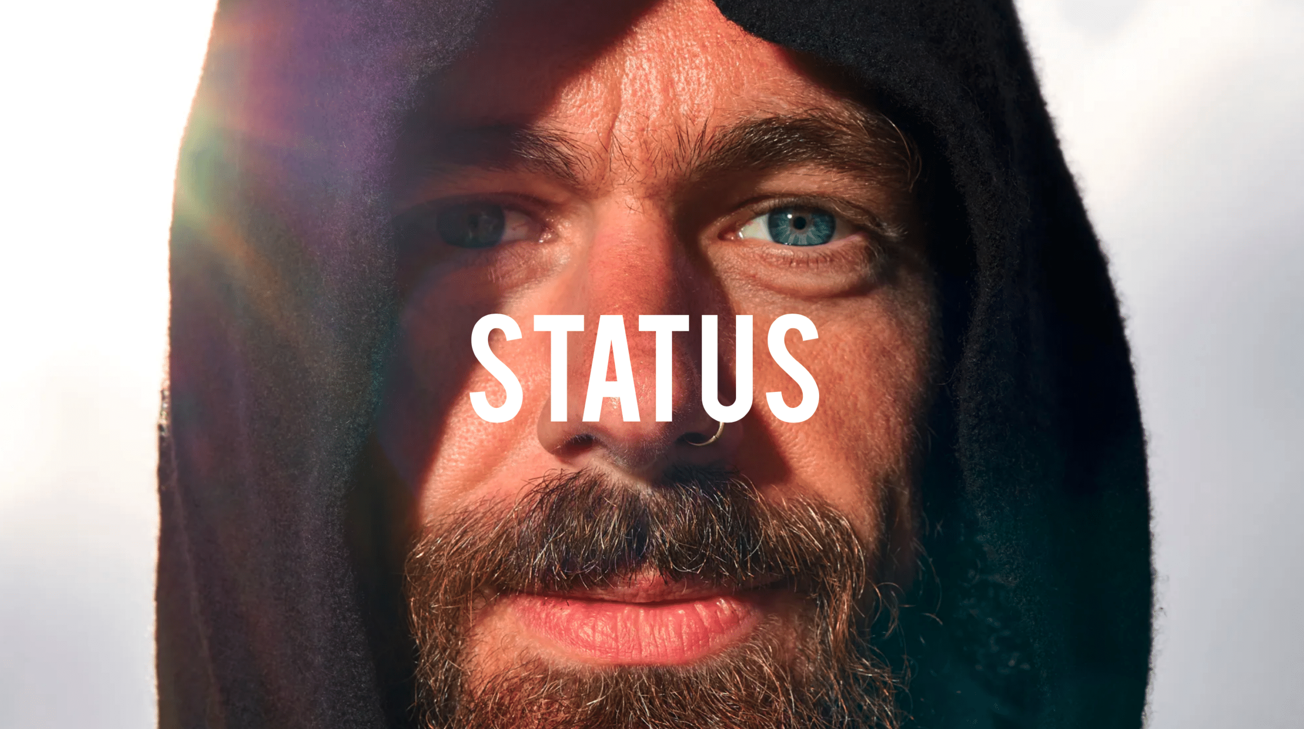 A close-up of a person with blue eyes, a beard, and a mustache, wearing a black hood. They are looking directly at the camera, with sunlight creating a slight lens flare on the left side of the image. The word "STATUS" is written across the center of the image. - Choice Hacking Academy A close-up of a person with blue eyes, a beard, and a mustache, wearing a black hood. They are looking directly at the camera, with sunlight creating a slight lens flare on the left side of the image. The word "STATUS" is written across the center of the image.