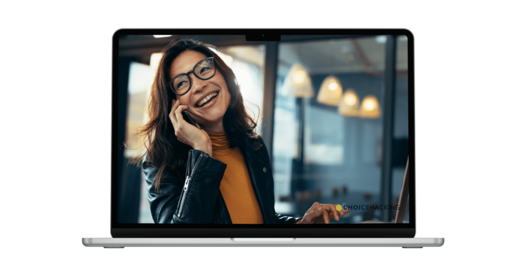 A woman with glasses and long hair is smiling while talking on a smartphone. She is wearing a yellow top and a leather jacket. The image is displayed on a laptop screen, with pendant lights and a window in the blurred background.