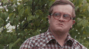 A person with large, round glasses and a serious expression is outdoors, standing in front of green foliage, wearing a checked shirt. The person turns their head to the side as if looking at something off-screen. - Choice Hacking Academy A person with large, round glasses and a serious expression is outdoors, standing in front of green foliage, wearing a checked shirt. The person turns their head to the side as if looking at something off-screen.