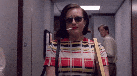A woman wearing sunglasses and a plaid dress confidently walks down an office hallway with a lit cigarette in her mouth. She is carrying wooden boards under her arm and has a determined expression while a man in the background turns to look at her.