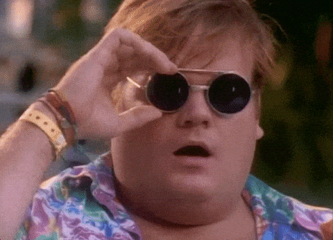 A surprised man wearing round black sunglasses and a colorful shirt lowers his glasses to look more closely, with his mouth open in shock or amazement.